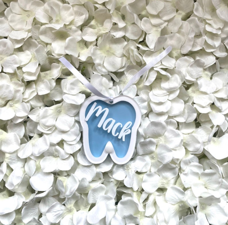 May include: A blue tooth-shaped ornament with the name "Mack" written in white on it. The ornament is hanging from a white ribbon and is surrounded by white flowers.
