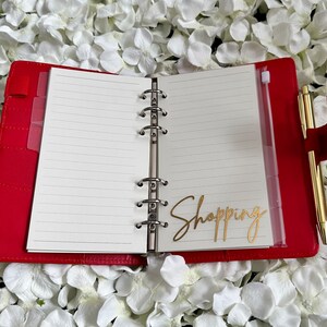 Puede incluir: Una agenda encuadernada en cuero rojo con un bol&iacute;grafo dorado y una pegatina dorada que dice "Shopping" en el interior.