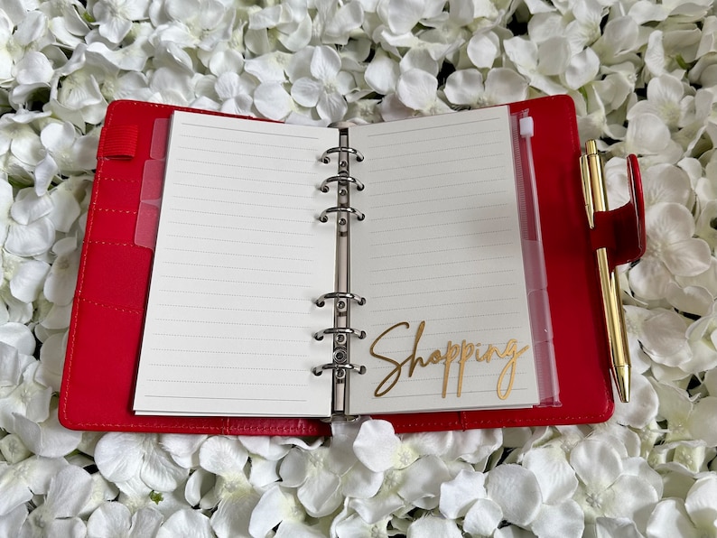 Puede incluir: Una agenda encuadernada en cuero rojo con un bol&iacute;grafo dorado y una pegatina dorada que dice "Shopping" en el interior.