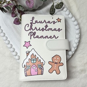 May include: White planner with a pink and white gingerbread house and gingerbread man design. The planner has the text "Laura's Christmas Planner" in pink lettering.