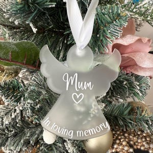 May include: A frosted glass angel Christmas ornament with the word "Mum" and a heart symbol, plus the phrase "In loving memory." The angel has outstretched wings and is suspended by a white ribbon.