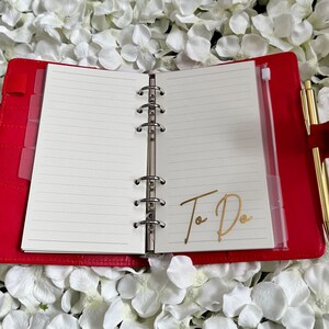Puede incluir: Planificador encuadernado en cuero rojo con un bolsillo con cremallera dorado y un bol&iacute;grafo dorado. El planificador tiene p&aacute;ginas rayadas y las palabras "To Do" escritas en dorado en la p&aacute;gina derecha.