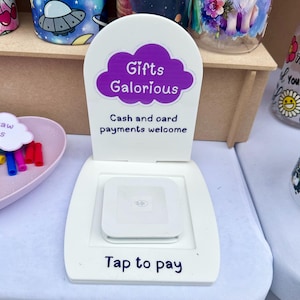 May include: A white stand with a purple cloud that says "Gifts Galorious" and "Cash and card payments welcome". A white square device with the text "Tap to pay" is on the stand.