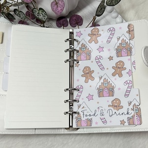 May include: A white planner with a gingerbread house and candy cane pattern on the tab dividers. The tab dividers are labeled "Food & Drink".