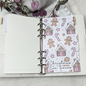 May include: A white planner with a gingerbread house and candy cane pattern on the tabs. The tabs are labeled "Christmas Eve Boxes".