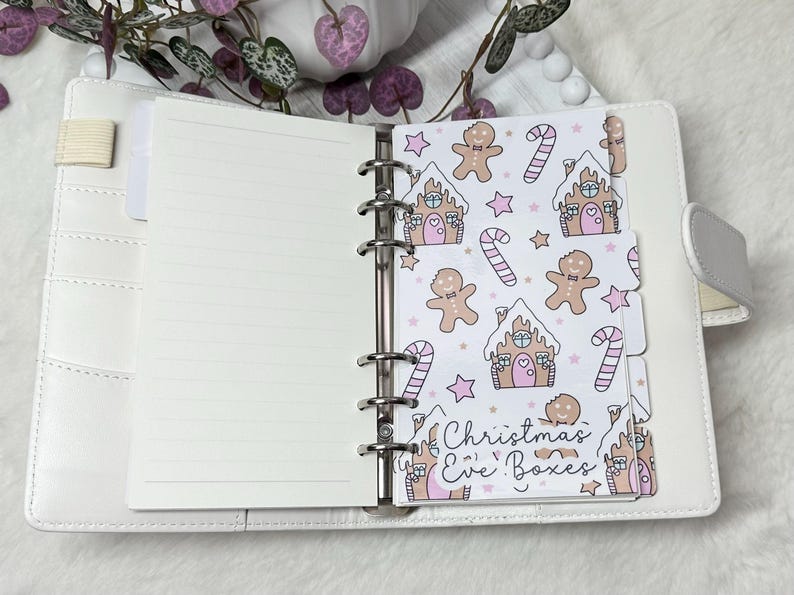 May include: A white planner with a gingerbread house and candy cane pattern on the tabs. The tabs are labeled "Christmas Eve Boxes".
