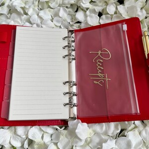 Puede incluir: Planificador encuadernado en cuero rojo con un bolsillo de pl&aacute;stico transparente etiquetado como "Recibos" y un bol&iacute;grafo dorado.