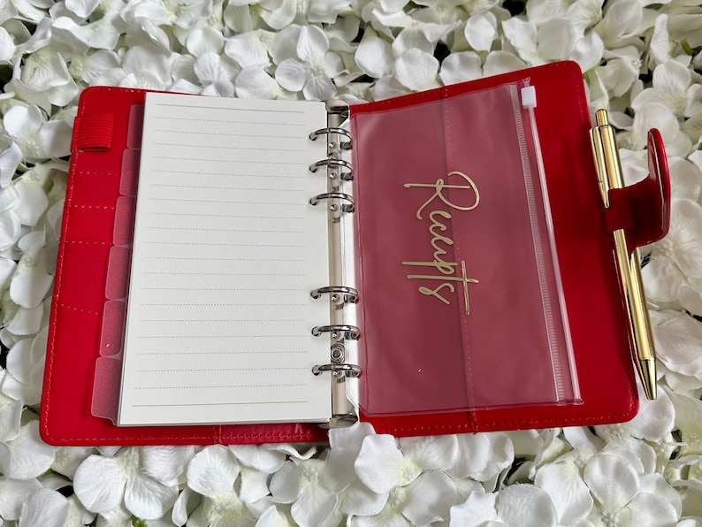 Puede incluir: Planificador encuadernado en cuero rojo con un bolsillo de pl&aacute;stico transparente etiquetado como "Recibos" y un bol&iacute;grafo dorado.