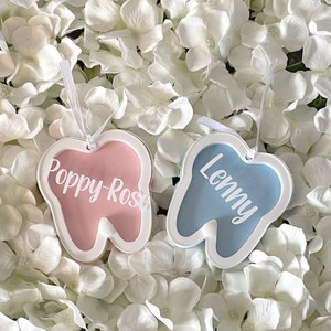 May include: Two tooth-shaped ornaments with white borders. The ornament on the left is pink with the name "Poppy-Rose" written in white. The ornament on the right is blue with the name "Lenny" written in white.