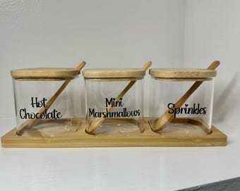 May include: A set of three glass jars with wooden lids and wooden spoons. The jars are labeled "Hot Chocolate", "Mini Marshmallows", and "Sprinkles".