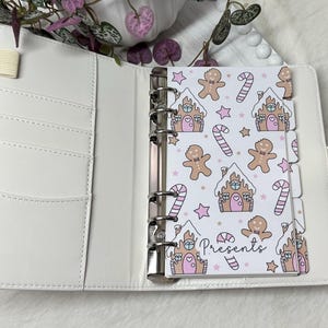 May include: White planner with a gingerbread house and gingerbread man pattern. The planner has a white ribbon bookmark and a silver ring binder. The planner has the word "Presents" written on it.