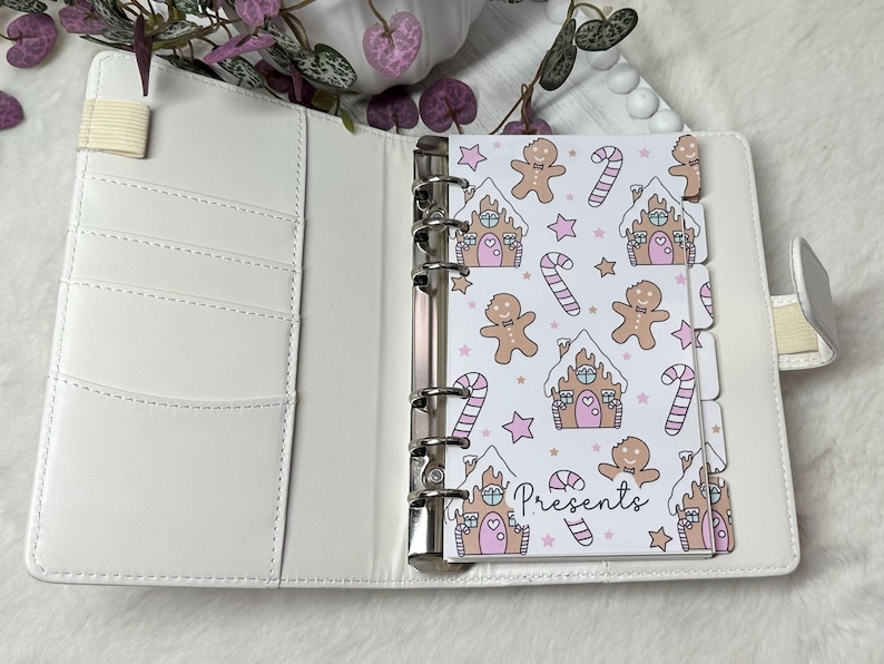 May include: White planner with a gingerbread house and gingerbread man pattern. The planner has a white ribbon bookmark and a silver ring binder. The planner has the word "Presents" written on it.