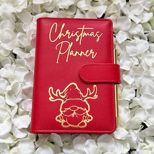 Puede incluir: Planificador de cuero rojo con una inscripci&oacute;n dorada que dice "Christmas Planner". Un gnomo dorado con cuernos de reno aparece en la portada.