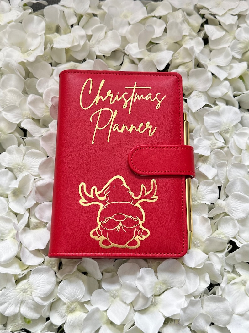 Puede incluir: Planificador de cuero rojo con una inscripci&oacute;n dorada que dice "Christmas Planner". Un gnomo dorado con cuernos de reno aparece en la portada.