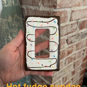 K&ouml;nnte beinhalten: Ein Lichtschalter-Abdeckung, die wie ein hei&szlig;er Schokoladen-Sundae bemalt ist. Die Abdeckung ist braun mit wei&szlig;em Frosting und bunten Streuseln.