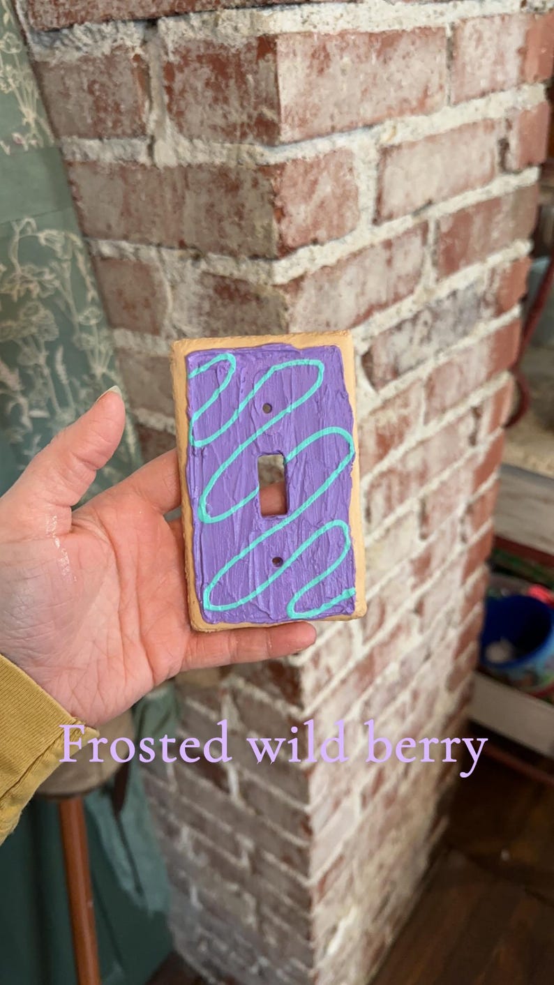 K&ouml;nnte beinhalten: Ein Lichtschalter-Abdeckung, die so bemalt ist, dass sie wie ein gefrosteter Wildbeere Pop-Tart aussieht. Die Abdeckung ist beige mit lila Frosting und einem hellblauen Wirbelmuster.