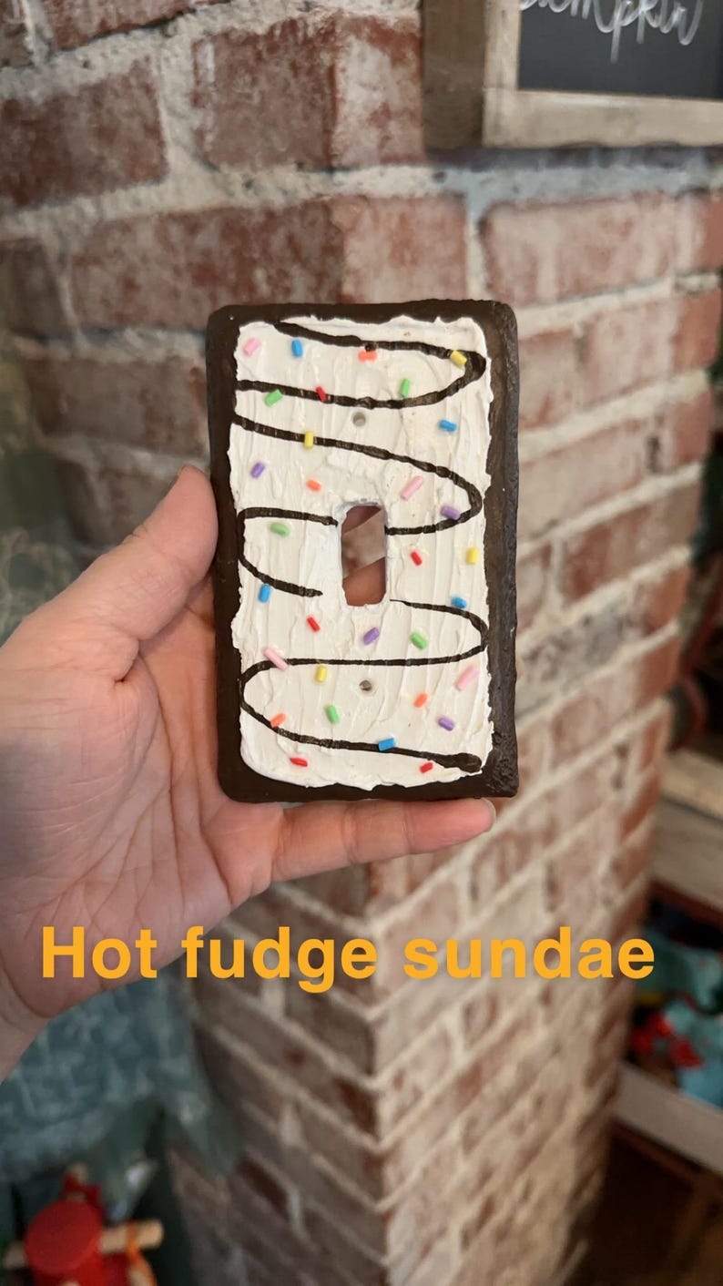K&ouml;nnte beinhalten: Ein Lichtschalter-Abdeckung, die wie ein hei&szlig;er Schokoladen-Sundae dekoriert ist. Die Abdeckung ist braun mit wei&szlig;em Frosting und Schokoladensauce und hat bunte Streusel.
