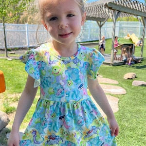 May include: A young girl wearing a blue and green dress with a pattern of characters from the movie Monsters Inc. The dress has a ruffled neckline and short sleeves. The girl is smiling and looking at the camera.