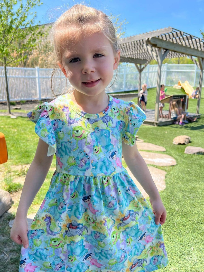 May include: A young girl wearing a blue and green dress with a pattern of characters from the movie Monsters Inc. The dress has a ruffled neckline and short sleeves. The girl is smiling and looking at the camera.