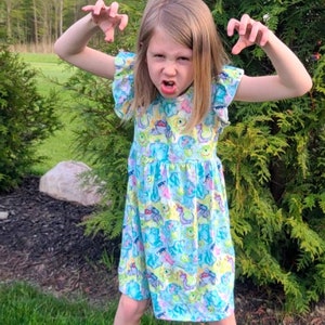 May include: A young girl wearing a blue dress with a colorful monster print. She is making a funny face and has her hands up in the air.
