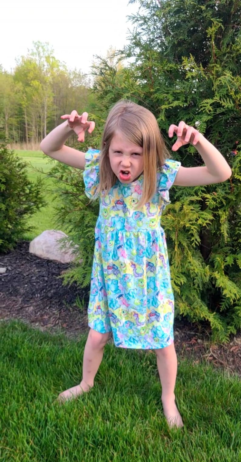 May include: A young girl wearing a blue dress with a colorful monster print. She is making a funny face and has her hands up in the air.
