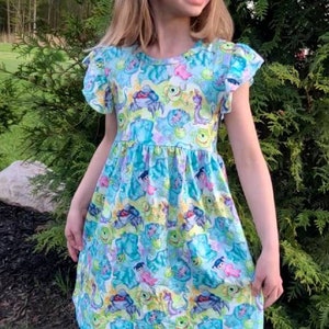 May include: A young girl wearing a blue and green dress with a pattern of cartoon monsters. The dress has short sleeves with ruffles and a gathered skirt. The girl is standing in a grassy area.