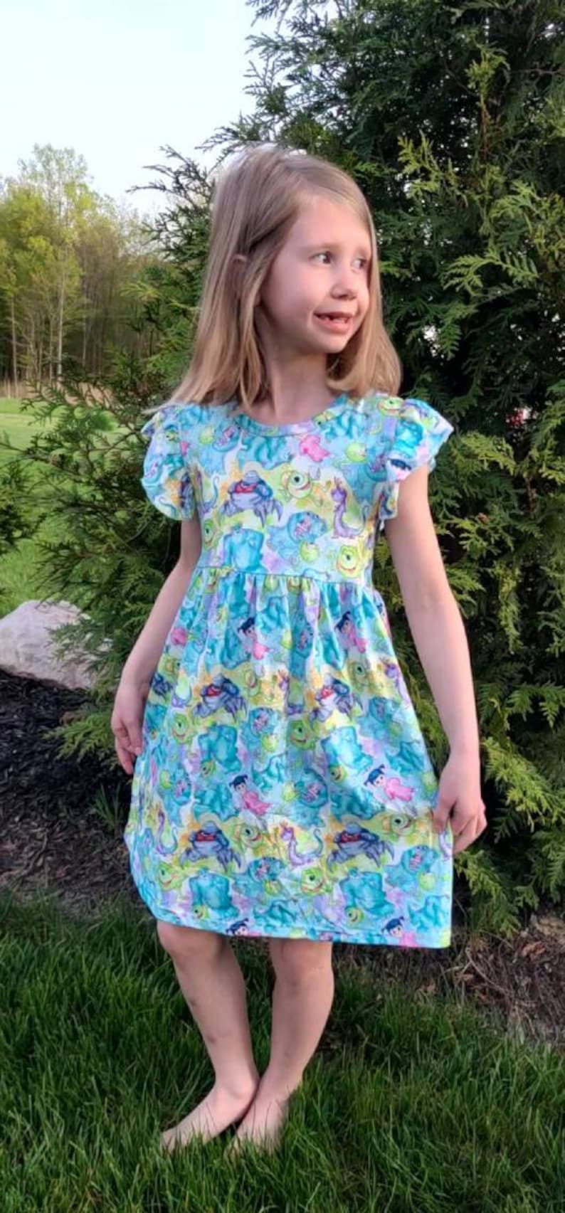 May include: A young girl wearing a blue and green dress with a pattern of cartoon monsters. The dress has short sleeves with ruffles and a gathered skirt. The girl is standing in a grassy area.
