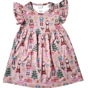 Puede incluir: Un vestido rosa sin mangas con un estampado festivo de Cascanueces. El vestido tiene un escote con volantes y mangas cortas con volantes. El estampado presenta soldados Cascanueces, árboles de Navidad y relojes.