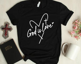 May include: Black t-shirt with the text "God is Love" in a white script font, featuring a heart and cross design. A wooden cross, a closed Holy Bible, and a white mug are also visible.