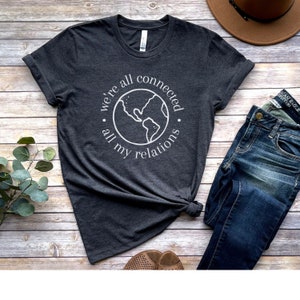 May include: Dark gray t-shirt with the text "we're all connected, all my relations" encircling a white outline of the Earth. The shirt is styled with blue jeans and a brown hat on a wooden surface.