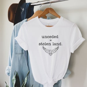 May include: White t-shirt with the text "unceded = stolen land" and a graphic of a bird with outstretched wings. The shirt is hanging on a wooden hanger with a denim shirt and a brown hat in the background.