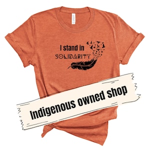 May include: Orange t-shirt with the text "I stand in SOLIDARITY" and a black feather design with birds. The shirt also has the text "Indigenous owned shop" on a white banner.