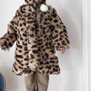 May include: A doll wearing a faux fur coat and matching hat with a leopard print pattern. The coat is brown and tan with dark brown spots. A white flower is pinned on the coat. The doll is also wearing brown pants and black shoes.