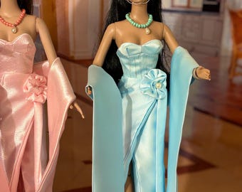 May include: Two fashion dolls wearing elegant gowns. The doll on the left is wearing a pink gown with a matching hat and a pearl necklace. The doll on the right is wearing a light blue gown with a matching hat and a turquoise necklace.