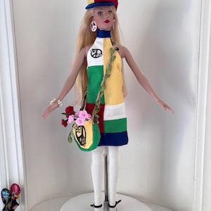 May include: A doll wearing a colorful, patchwork dress with a peace sign. The doll has long blonde hair, a matching hat, and a small purse with pink roses. The doll is wearing white stockings and black heels.