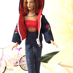 May include: A doll wearing a blue denim jacket with a red hood, a red crop top, blue denim pants, and a pearl necklace. The doll is holding a red purse and standing next to a pink bicycle.