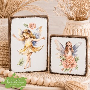 May include: Two rectangular wooden signs with distressed dark edges and white faces. The taller sign features a blonde fairy in a yellow dress with blue wings. The shorter sign shows a fairy with brown hair and blue wings surrounded by pink flowers.
