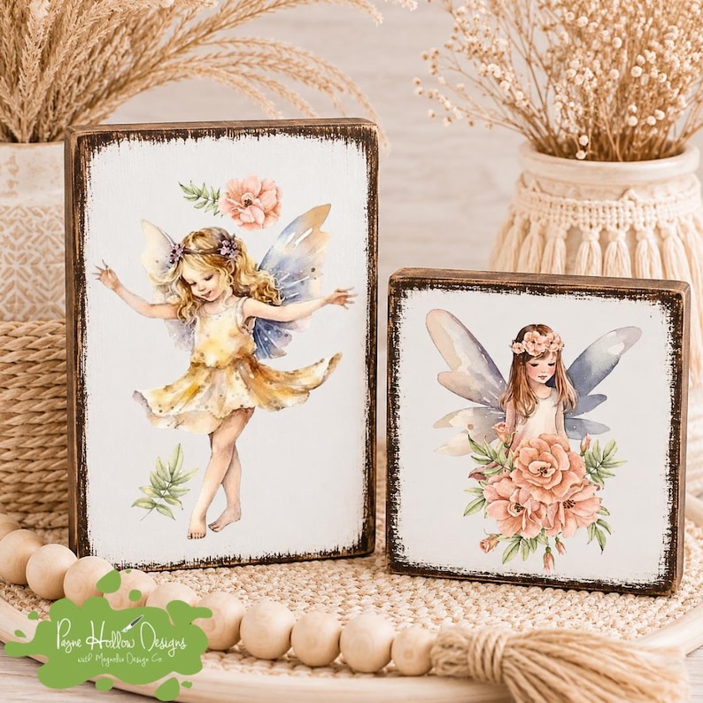 May include: Two rectangular wooden signs with distressed dark edges and white faces. The taller sign features a blonde fairy in a yellow dress with blue wings. The shorter sign shows a fairy with brown hair and blue wings surrounded by pink flowers.