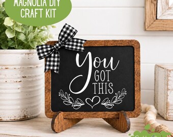 May include: A rectangular chalkboard with a dark brown wooden frame and a black and white buffalo check ribbon bow. The chalkboard features white text that reads You Got This with a heart and leaf graphic design. It rests on a wooden stand.