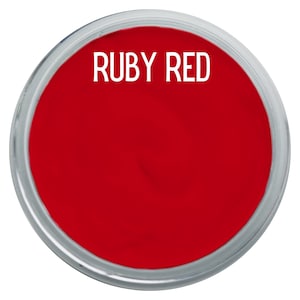 May include: A circular container filled with vibrant ruby red pigment or paint. The container features a clear, transparent rim and a smooth, solid red surface. The text "RUBY RED" is printed in a clean, white, sans-serif font across the top.