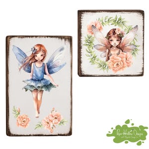 May include: Two rectangular wooden signs featuring watercolor-style illustrations of fairies. One sign shows a standing fairy in a blue dress with blue wings and peach flowers. The other shows a fairy with peach wings inside a green leaf wreath.