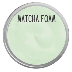 May include: A circular container filled with a smooth, light green substance. The text "MATCHA FOAM" is printed in black, sans-serif capital letters across the upper portion of the pale green surface, enclosed by a thin, metallic silver rim.