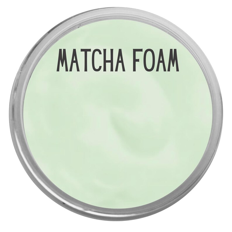 May include: A circular container filled with a smooth, light green substance. The text "MATCHA FOAM" is printed in black, sans-serif capital letters across the upper portion of the pale green surface, enclosed by a thin, metallic silver rim.