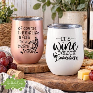 K&ouml;nnte beinhalten: Zwei stiellose Edelstahlbecher mit Deckeln. Der ros&eacute;goldene Becher tr&auml;gt den schwarzen Schriftzug of course I drink like a fish I'm a MERMAID mit einer Meerjungfrauenflosse. Der wei&szlig;e Becher tr&auml;gt den schwarzen Schriftzug IT'S wine O'CLOCK somewhere mit einem Weinglas-Symbol.