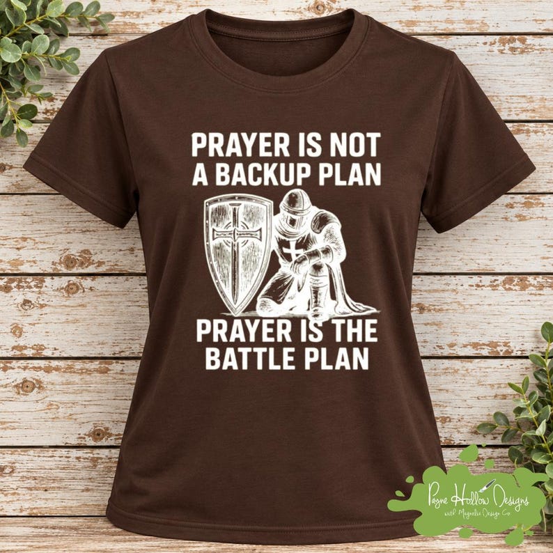 May include: A dark brown short-sleeve t-shirt featuring a white graphic of a knight kneeling with a shield. The text reads: PRAYER IS NOT A BACKUP PLAN PRAYER IS THE BATTLE PLAN. The design is centered on the chest of the shirt.