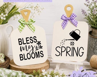 May include: Two white wooden signs with handles, one featuring a green gingham bow and the text BLESS MY BLOOMS, the other with a purple gingham bow and the text SPRING with a watering can graphic. The signs are displayed on small wooden stands.