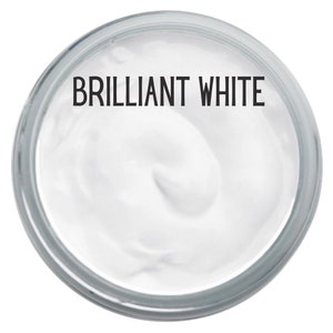 May include: A circular container filled with a thick, smooth, opaque white substance. The text BRILLIANT WHITE is printed in a bold, black, sans-serif font across the top portion of the white surface, centered within the clear glass rim.