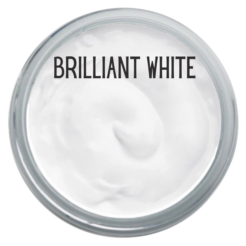 May include: A circular container filled with a thick, smooth, opaque white substance. The text BRILLIANT WHITE is printed in a bold, black, sans-serif font across the top portion of the white surface, centered within the clear glass rim.