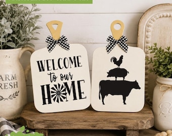May include: Two wooden cutting board signs with handles and black and white gingham bows. One sign features the black text "WELCOME to our HOME" with a windmill graphic. The other sign displays a black silhouette of a rooster, pig, and cow.