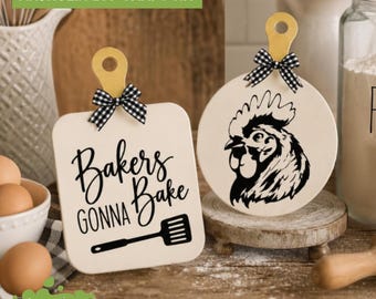 May include: Two small wooden cutting boards with handles and black and white gingham bows. One square board features the black text Bakers GONNA Bake and a spatula icon. The other round board displays a black graphic illustration of a rooster head.
